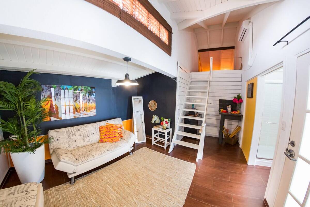 A modern living room with a white sofa, a large potted plant, and a staircase leading to an upper level.