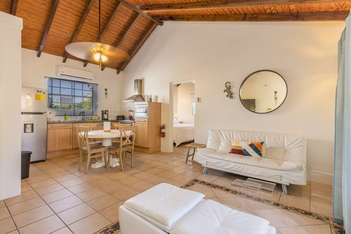 A spacious living area with a white sofa, a dining table with chairs, a kitchen with wooden cabinets, and a ceiling fan.