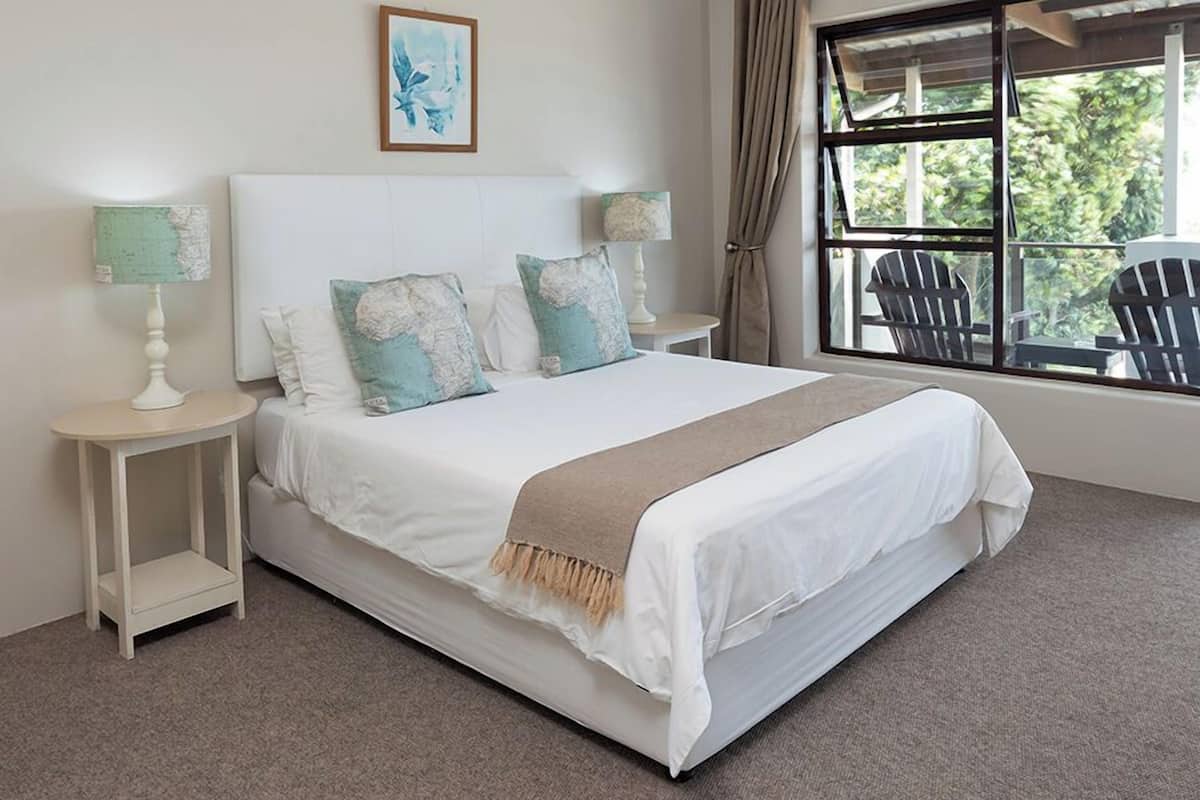 A bedroom with a large bed, two bedside tables with lamps, a window with curtains, and a view of greenery outside.