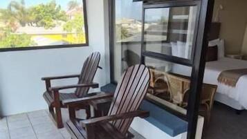 A balcony with two wooden chairs and a small table, a sliding glass door leading to a bedroom, and a view of the outdoors.