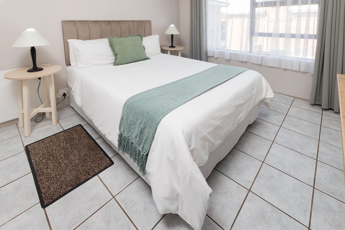 A neatly made bed with white bedding and a green throw, two bedside lamps, a small round table, and a view of a building through a window with curtains.