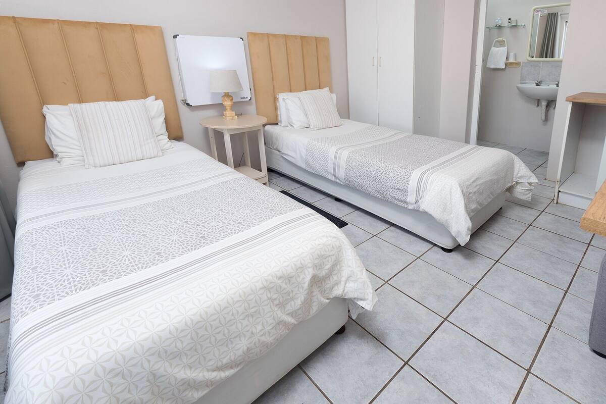 A hotel room with two beds, a small bedside table with a lamp, a whiteboard, a wardrobe, and a bathroom with a sink and mirror.