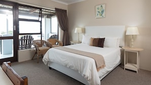 A hotel room with a large bed, two bedside tables with lamps, a wicker chair, and a view through a glass door. - Greystone Guest House (Jeffreys Bay)