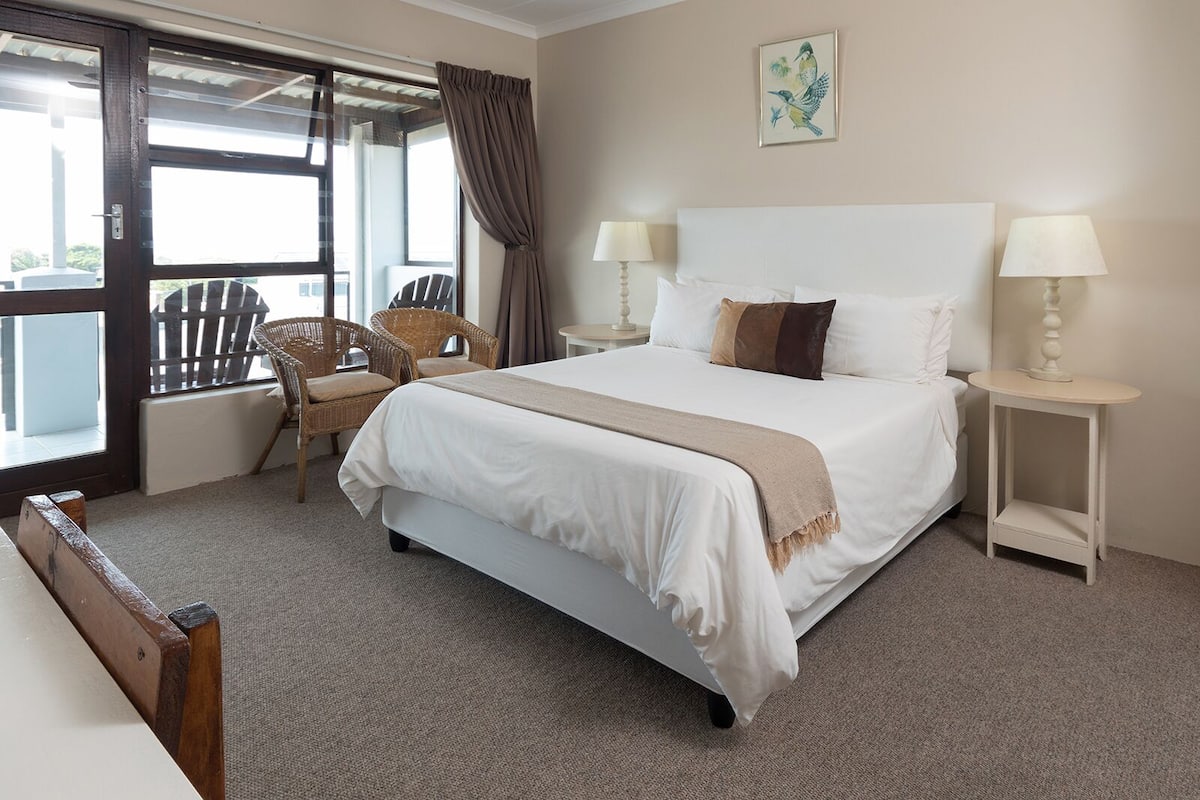 A hotel room with a large bed, two bedside tables with lamps, a wicker chair, and a view through a glass door.