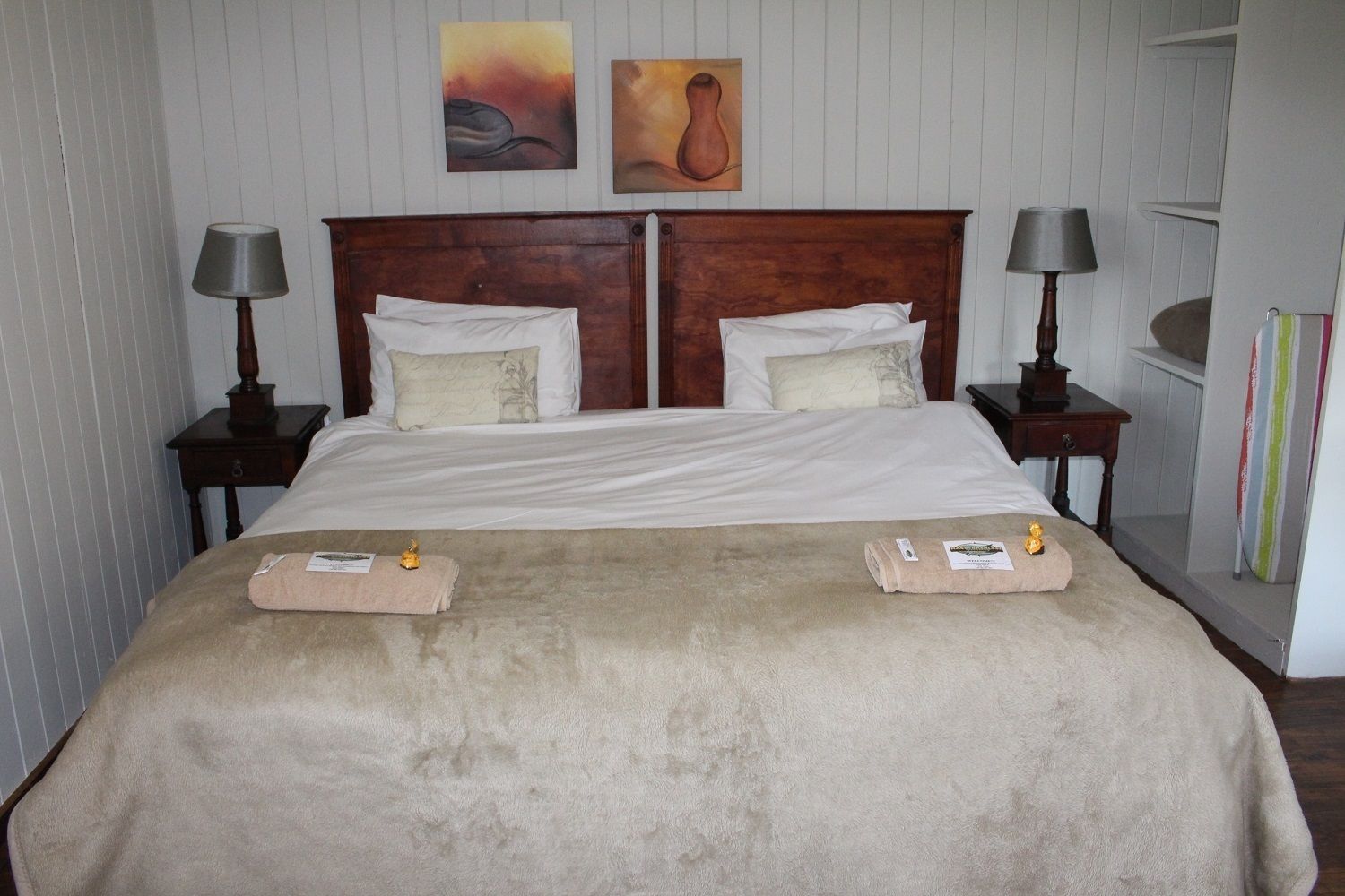 A double bed with a beige blanket, two bedside tables with lamps, and two paintings on the wall.