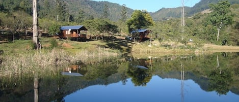 A cabin with a deck, surrounded by a natural landscape with hills and a calm lake reflecting the scenery.