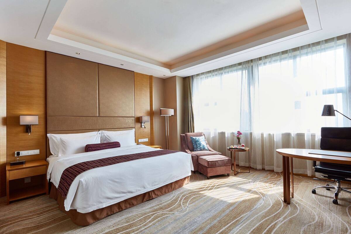 A hotel room with a large bed, a desk, a chair, and a small table with a lamp.