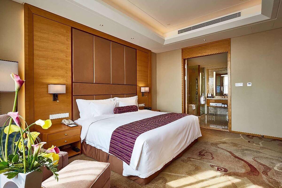 A hotel room with a large bed, bedside tables, a nightstand, a lamp, a vase with flowers, and a mirror.
