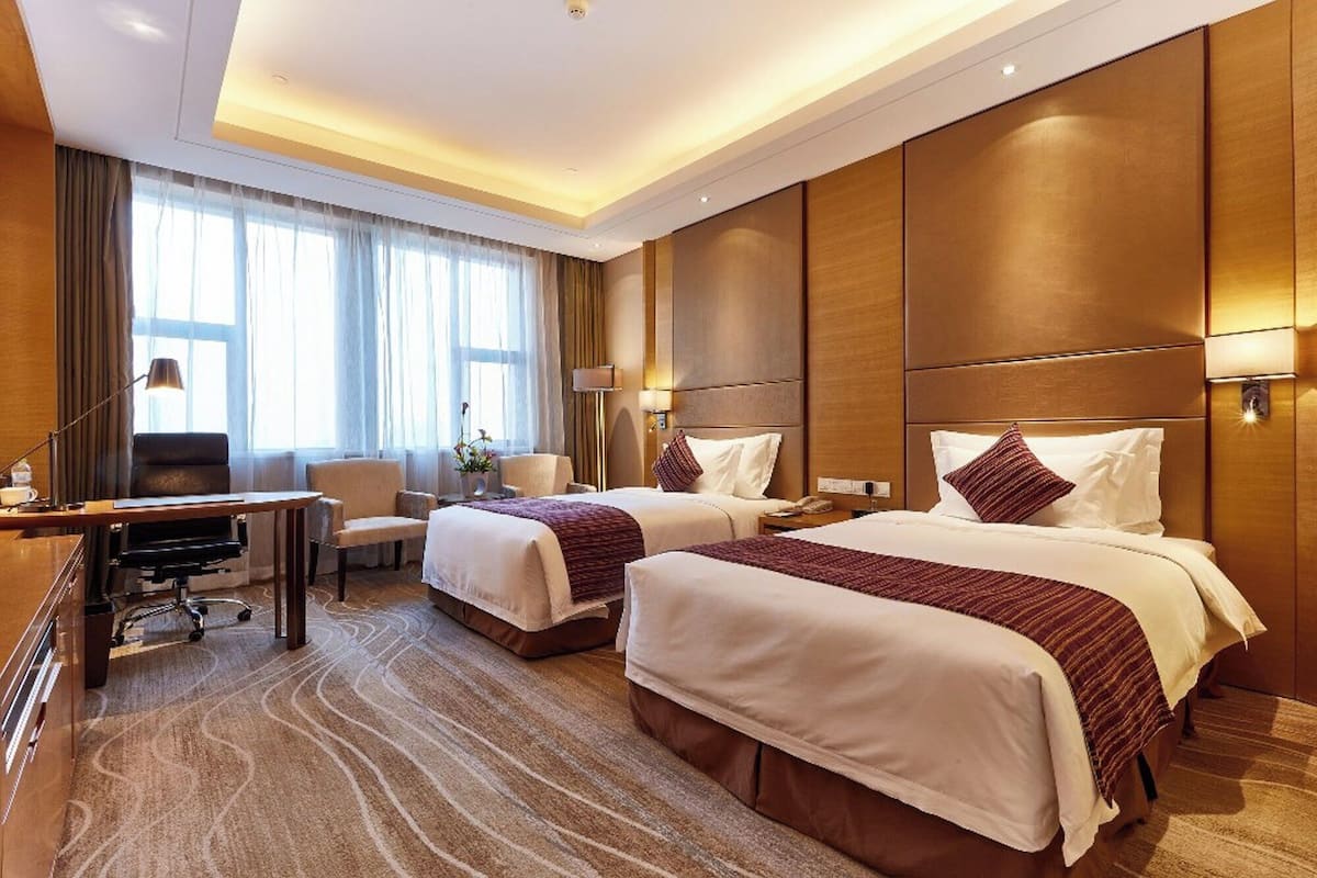 A hotel room with two beds, a desk, a chair, and a large window with curtains.