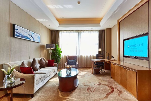 Habitación de hotel con sofá, mesa de centro, televisor y un ventanal con cortinas.