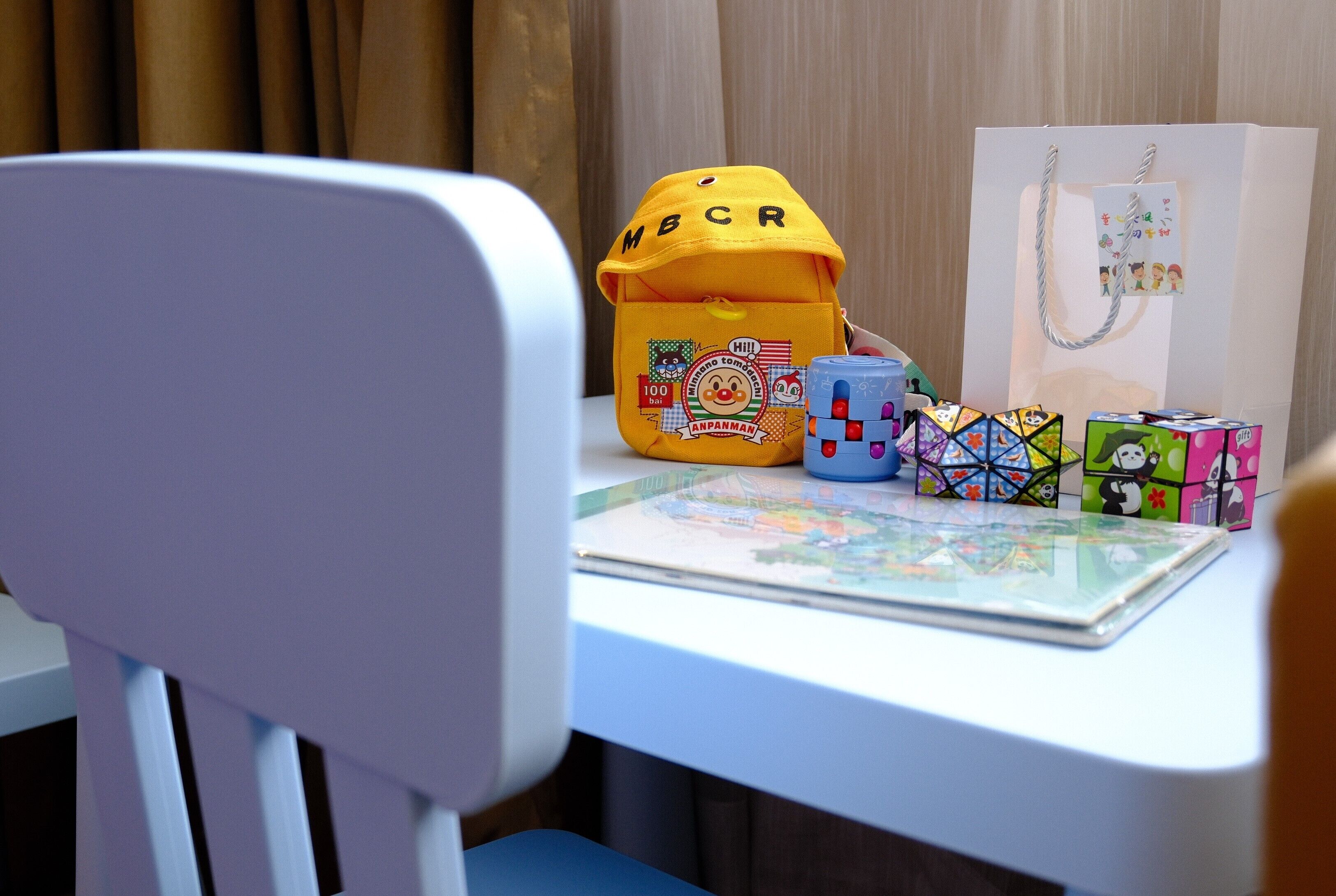A white chair with a table in front of it. On the table, there is a yellow bag with the letters "MBCR" on it, a white paper bag with a picture of children on it, and several colorful puzzle cubes.