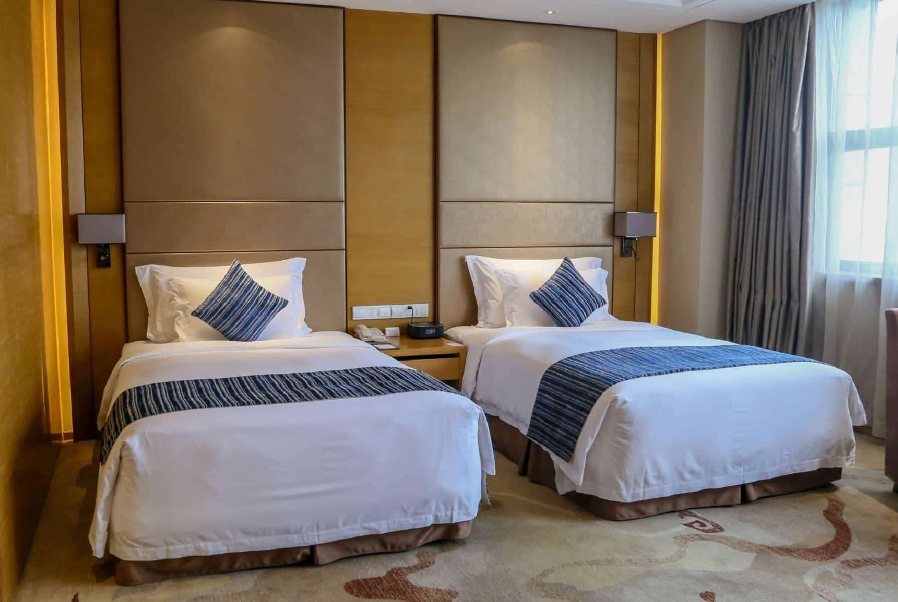 A hotel room with two beds, each with white bedding and blue accents, a wooden headboard, a nightstand with a lamp, and a large window with curtains.