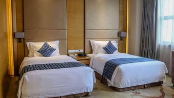 A hotel room with two beds, each with white bedding and blue accents, a wooden headboard, a nightstand with a lamp, and a large window with curtains.