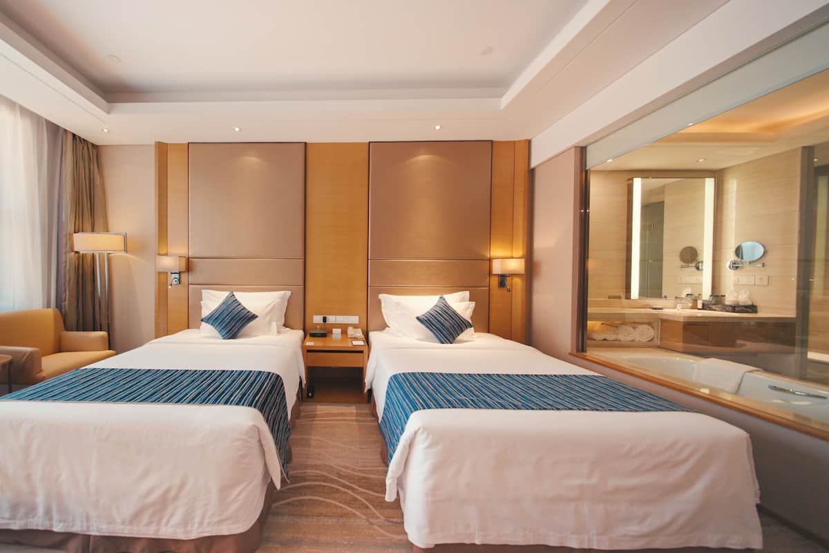 A hotel room with two beds, a sitting area with a chair and a table, a large mirror, and a view of the bathroom.