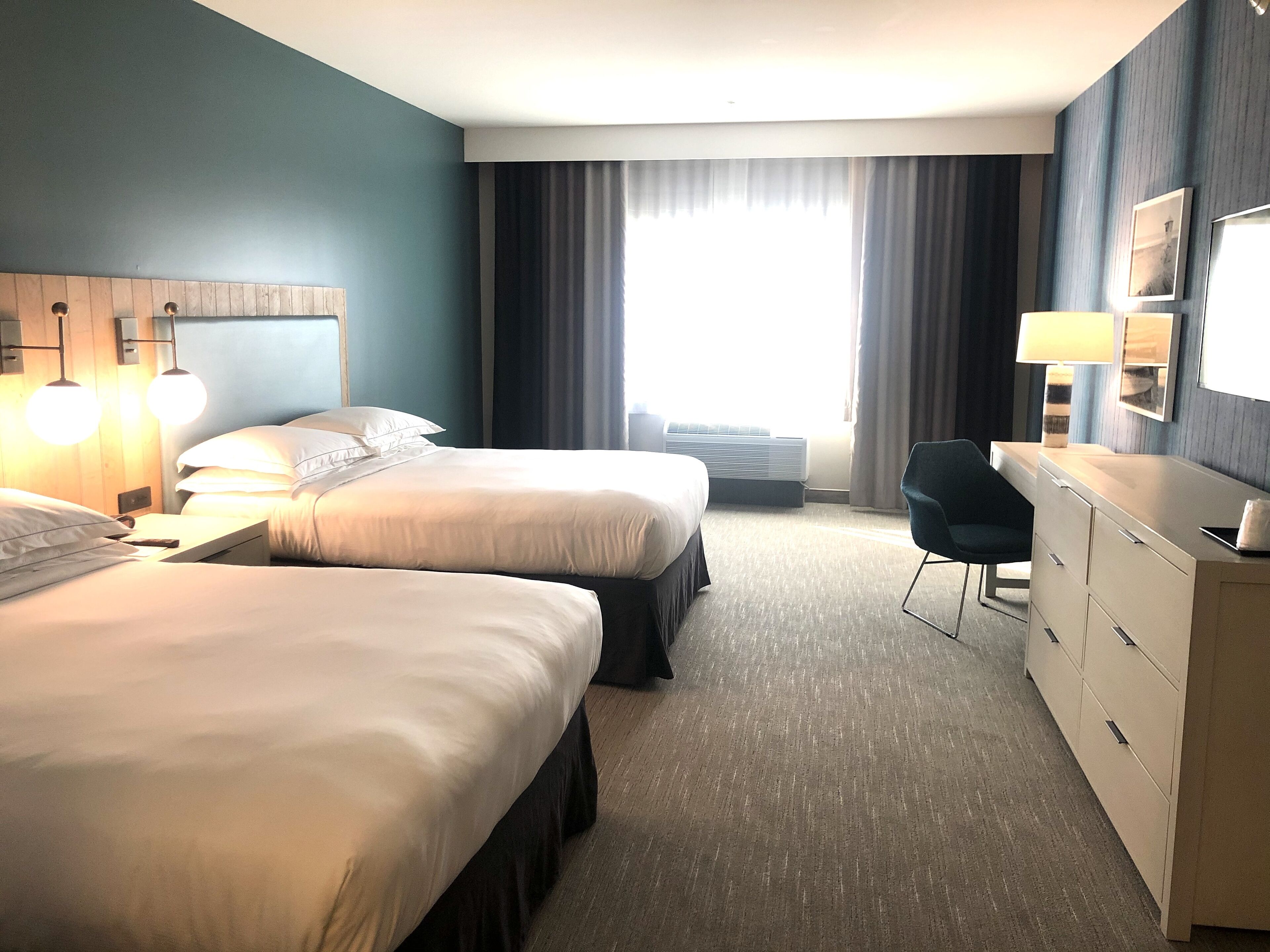 A hotel room with two beds, a desk, a chair, a dresser, and a large window with curtains.