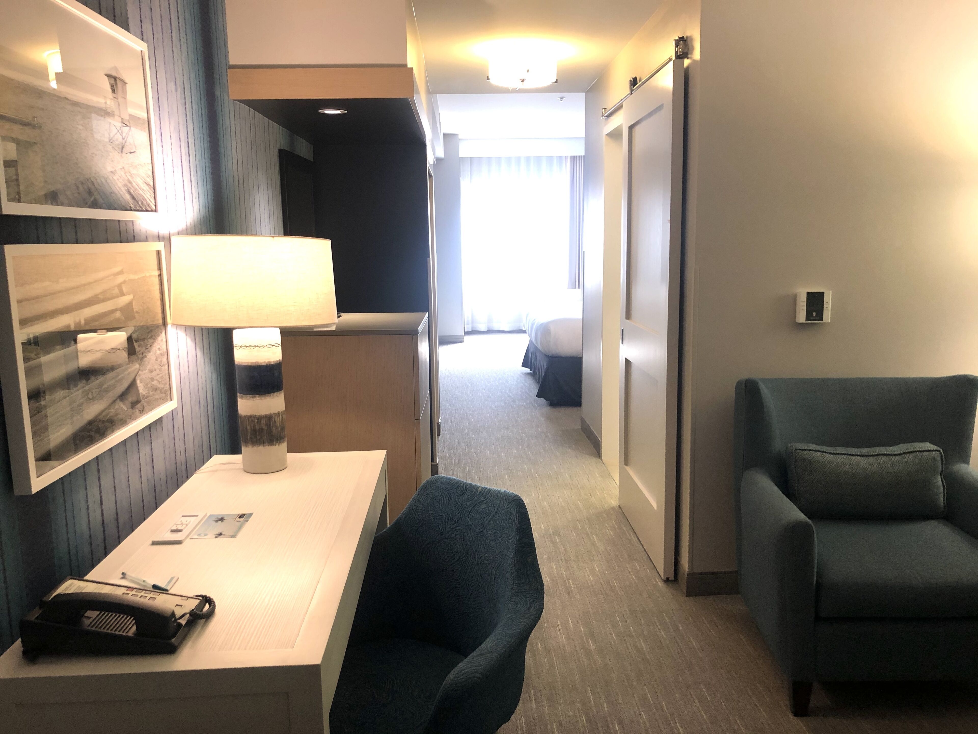 Ein Hotelzimmer mit Schreibtisch, Stuhl, Lampe und Blick auf den zum Schlafzimmer führenden Flur.