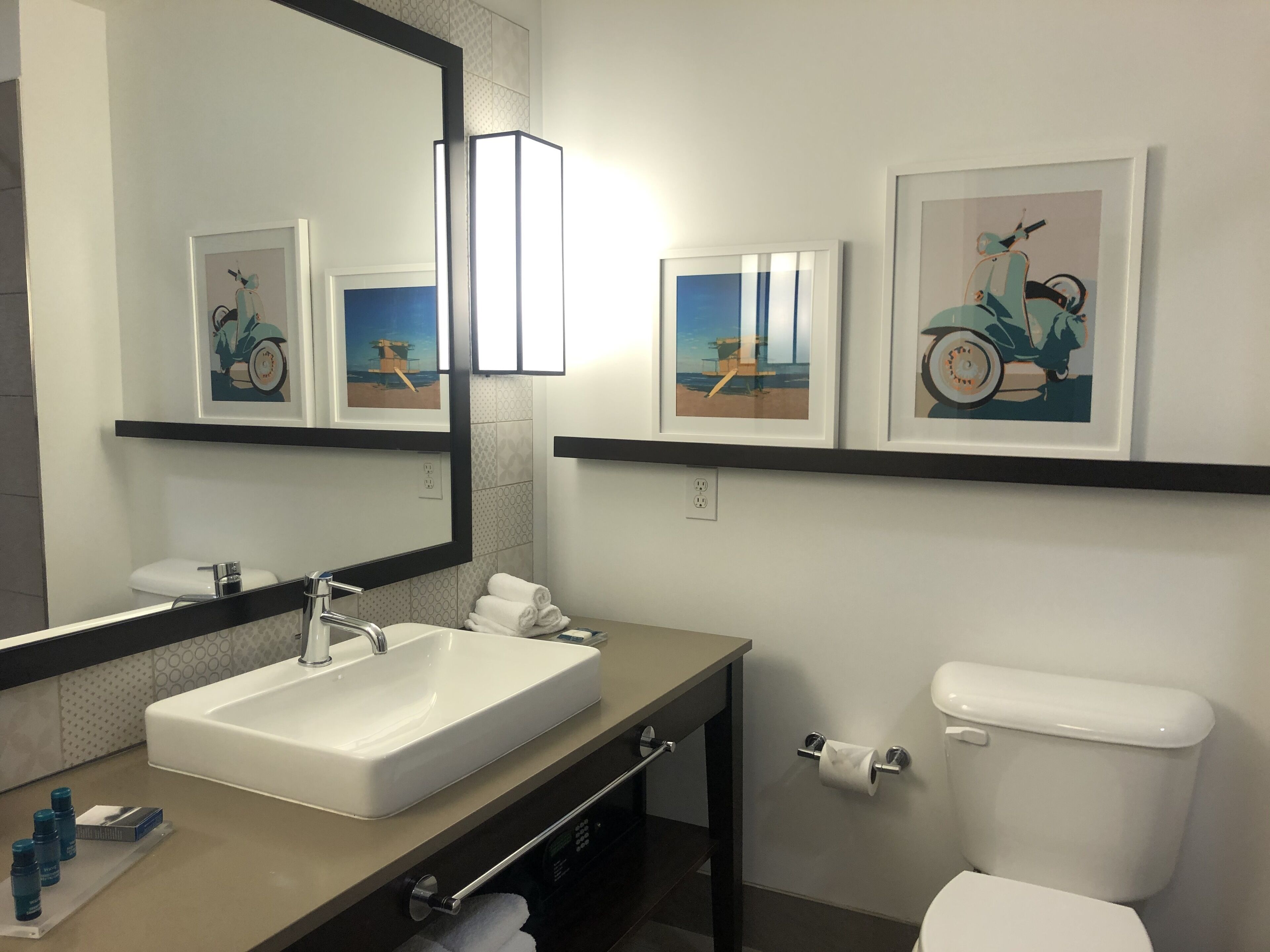 Un baño moderno con un espejo grande, un lavabo, un inodoro y obras de arte enmarcadas en las paredes.