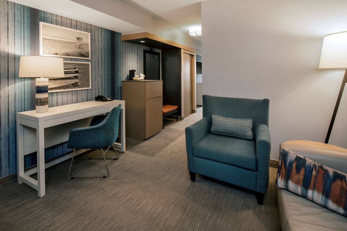 A hotel room with a desk, a blue armchair, a sofa, a lamp, and a bookshelf.