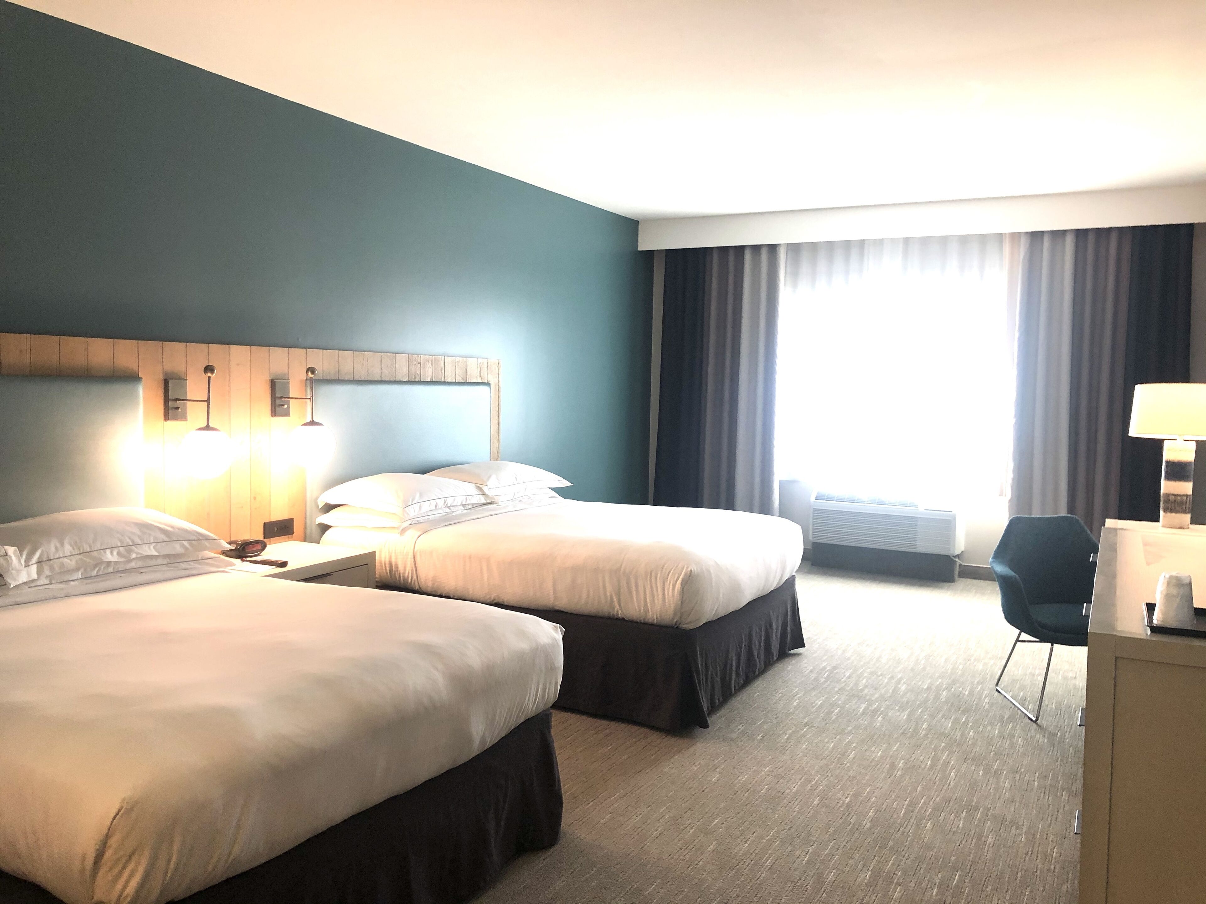 A hotel room with two beds, a desk, a chair, and a large window with curtains.