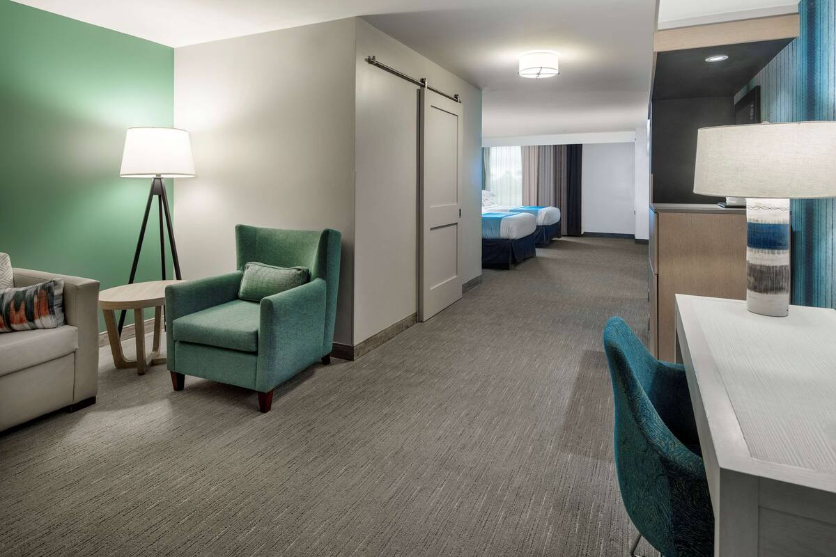 A hotel room with a green armchair, a desk, a lamp, a bed, and a wardrobe.