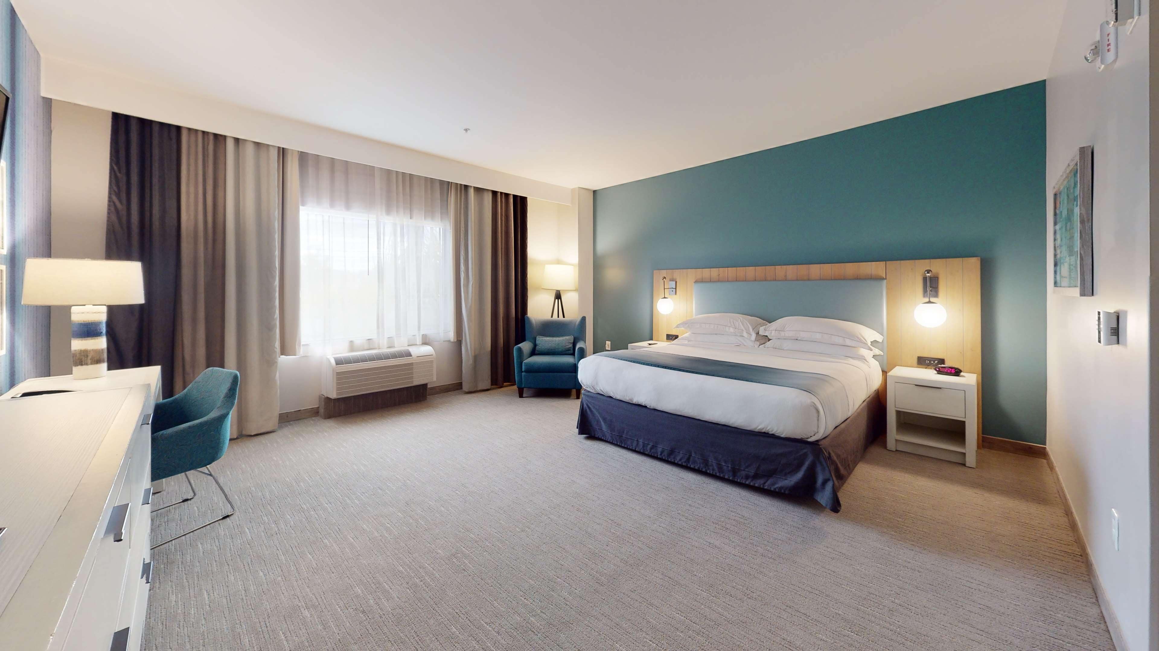 Habitación de hotel con cama, mesitas de noche, escritorio con lámpara, silla, ventana con cortinas y aire acondicionado de pared.
