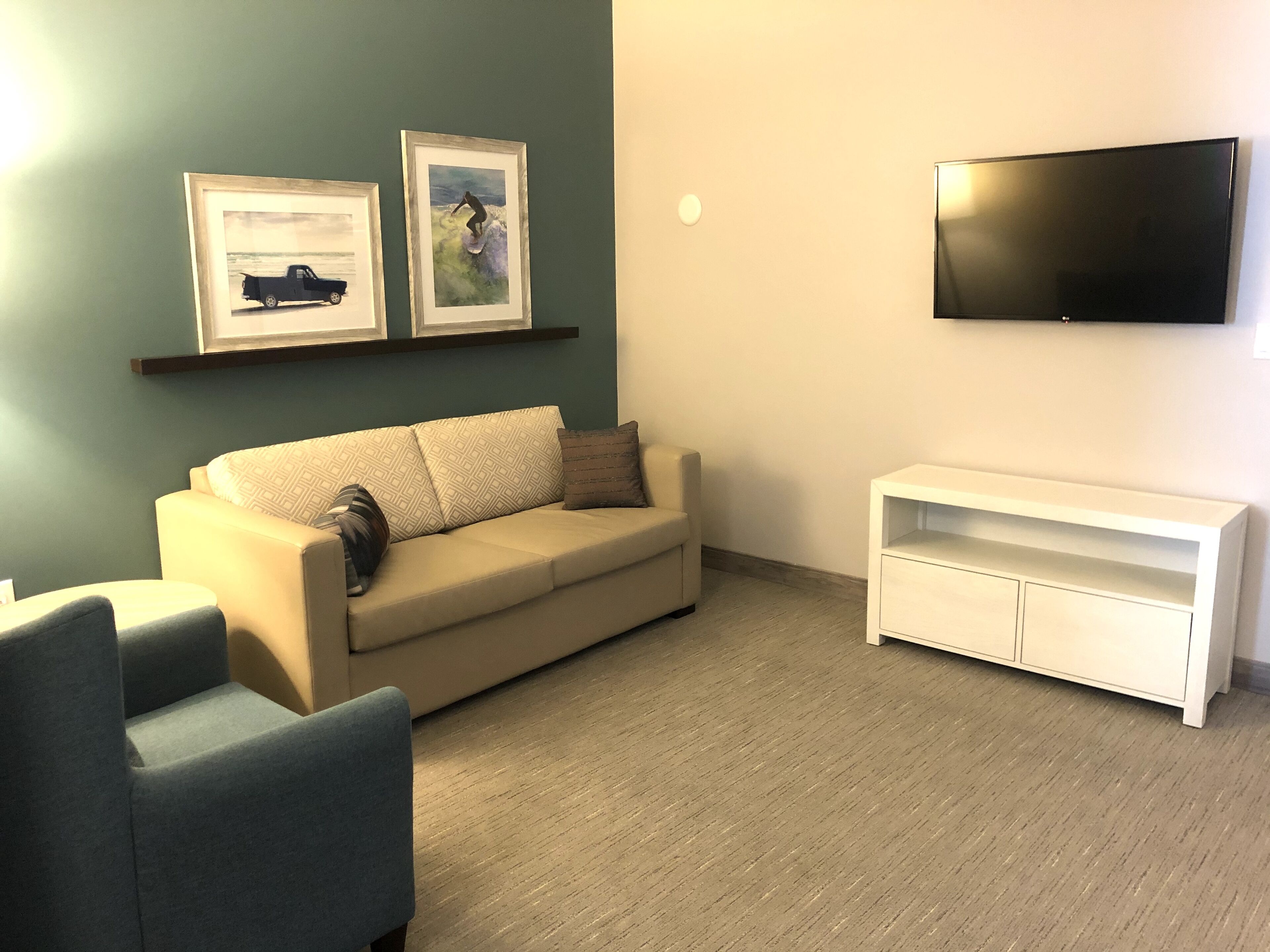 A living room with a beige sofa, a blue armchair, a white TV stand, and two framed artworks on the wall.