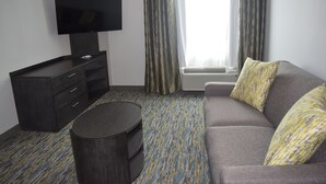 Ein modernes Hotelzimmer mit grauem Sofa, einem Flachbildfernseher auf einem dunklen Holzschrank, einem runden Beistelltisch und einem gemusterten Teppich.