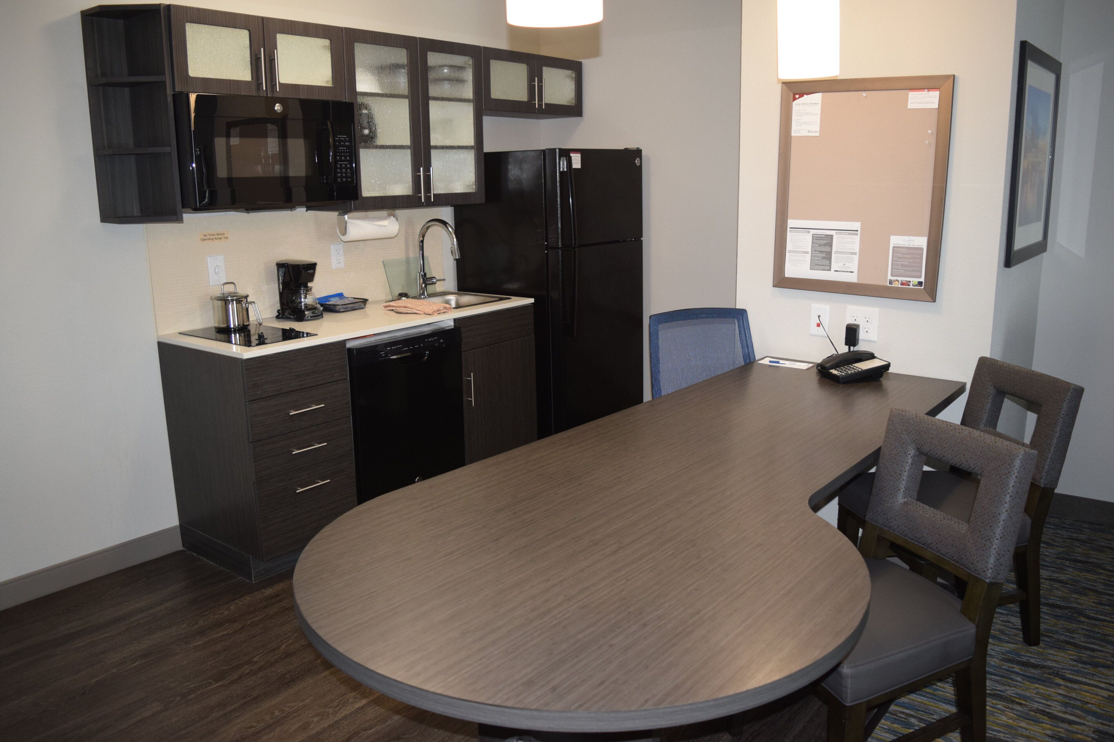 A compact kitchen with a microwave, coffee maker, and refrigerator. There's a dining table with chairs and a small conference table with a phone.