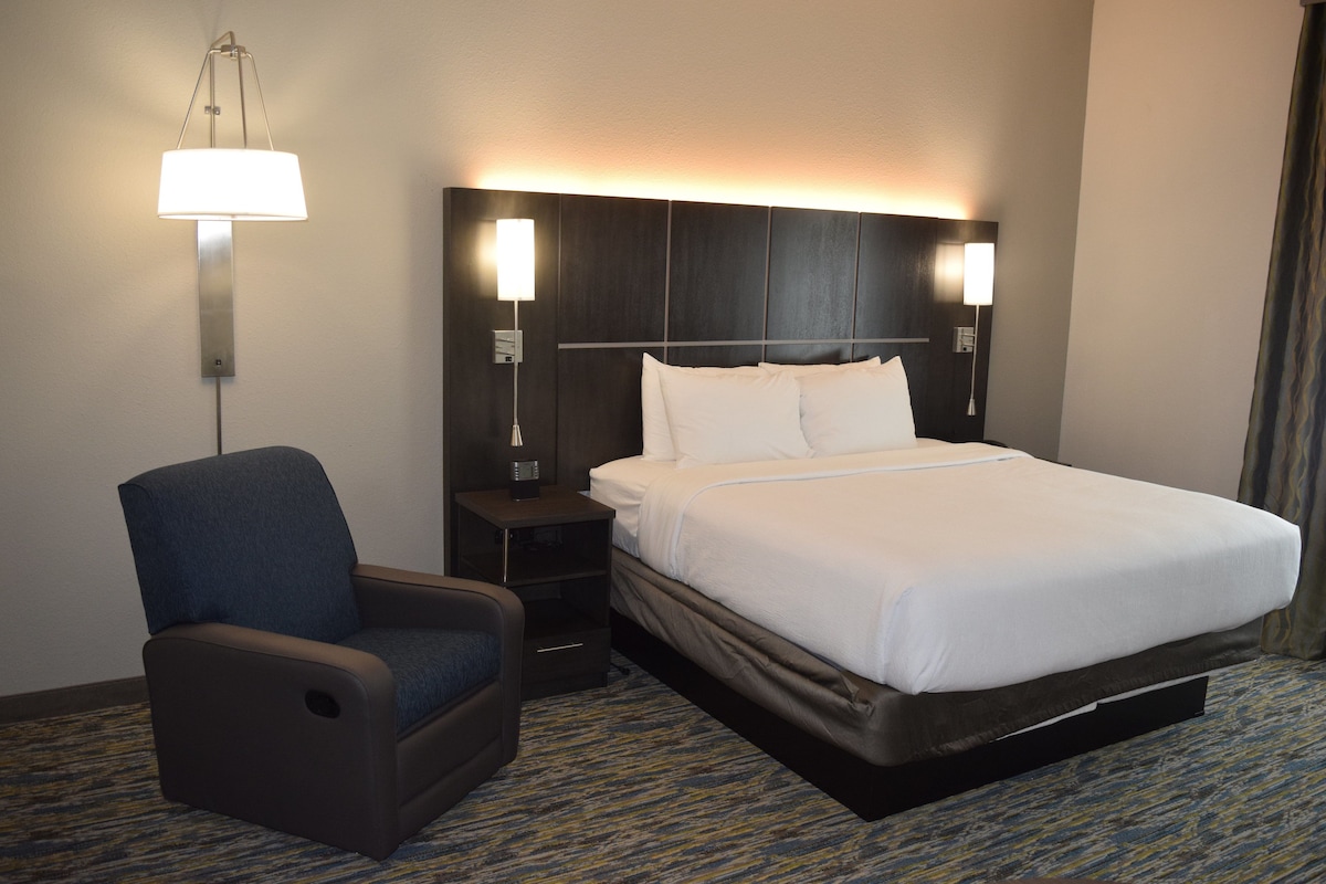Quarto de hotel com cama, poltrona, mesa de cabeceira e luminárias fixadas na parede.