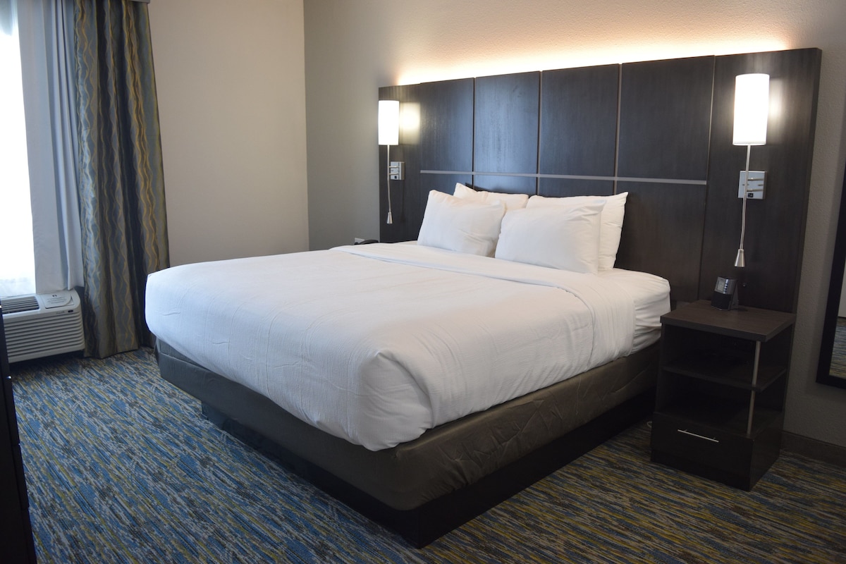 Quarto de hotel com uma cama grande, duas mesas de cabeceira e uma luminária de parede.