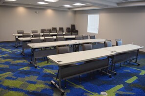 Un salón de conferencias con varias mesas y sillas, piso alfombrado y una ventana con persianas.