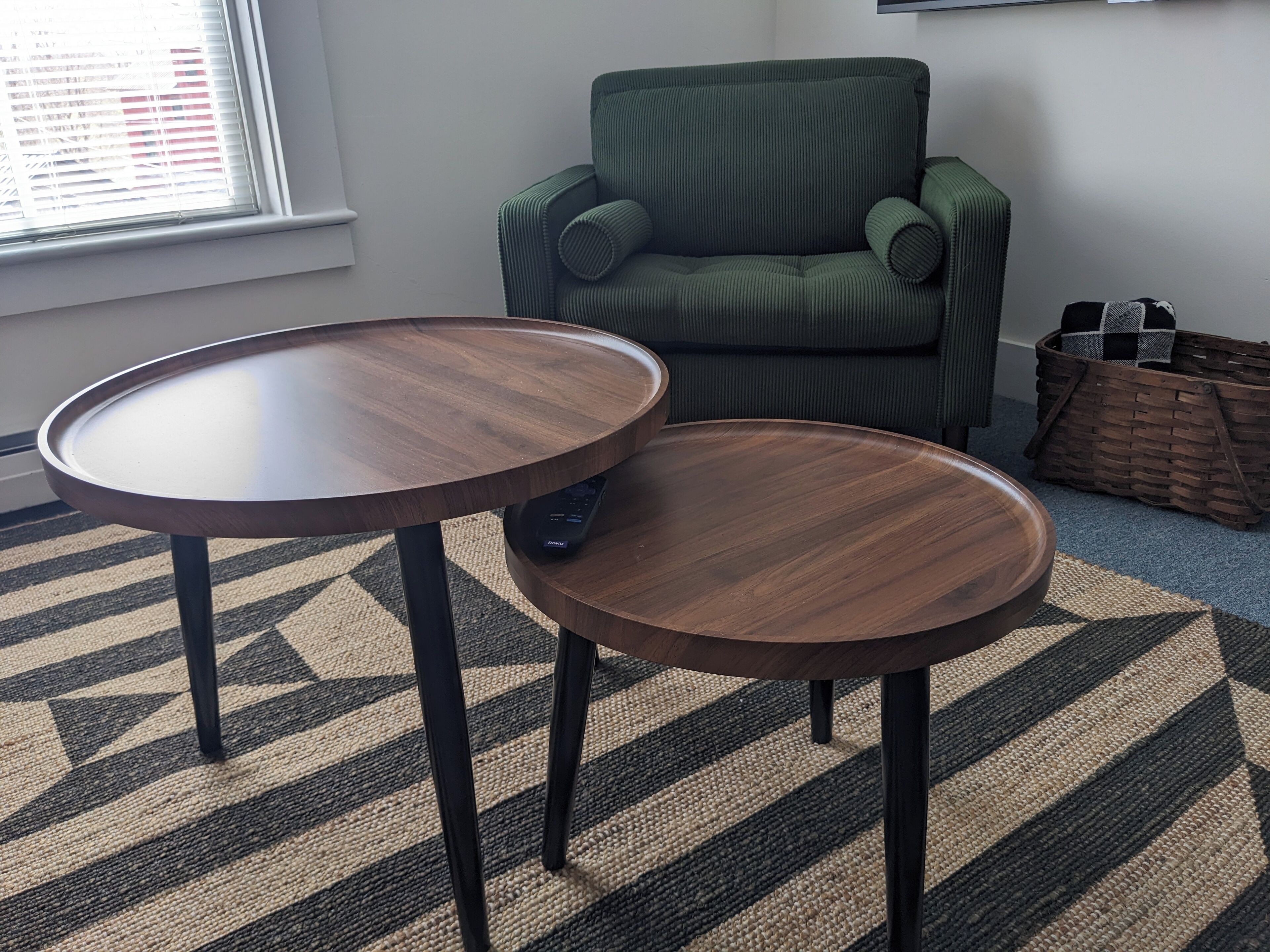 A room with two round wooden tables, a green armchair, a woven basket, and a patterned rug.