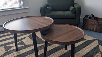 A room with two round wooden tables, a green armchair, a woven basket, and a patterned rug.