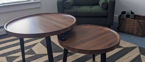 Een kamer met twee ronde houten tafels, een groene fauteuil, een gevlochten mand en een kamerkleed met dessin.