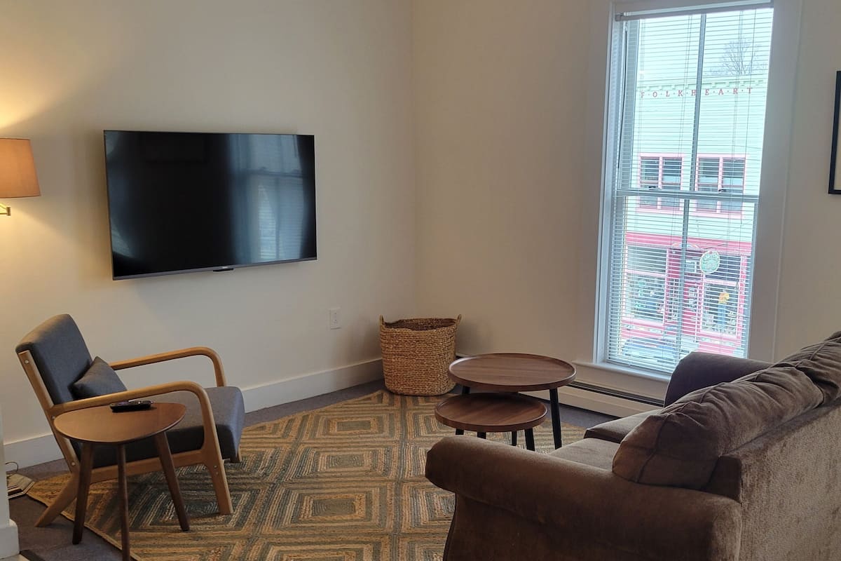 A living room with a flat-screen TV mounted on the wall, a sofa, a chair, two small tables, and a patterned rug.