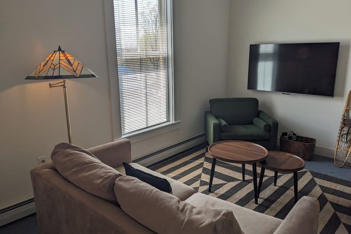 A living room with a sofa, a green armchair, a coffee table, a lamp, and a TV on the wall.