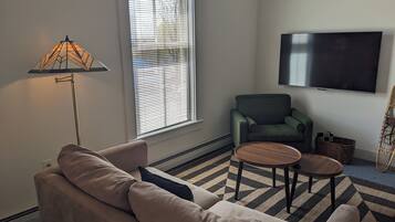 A living room with a sofa, a green armchair, a coffee table, a lamp, and a TV on the wall.