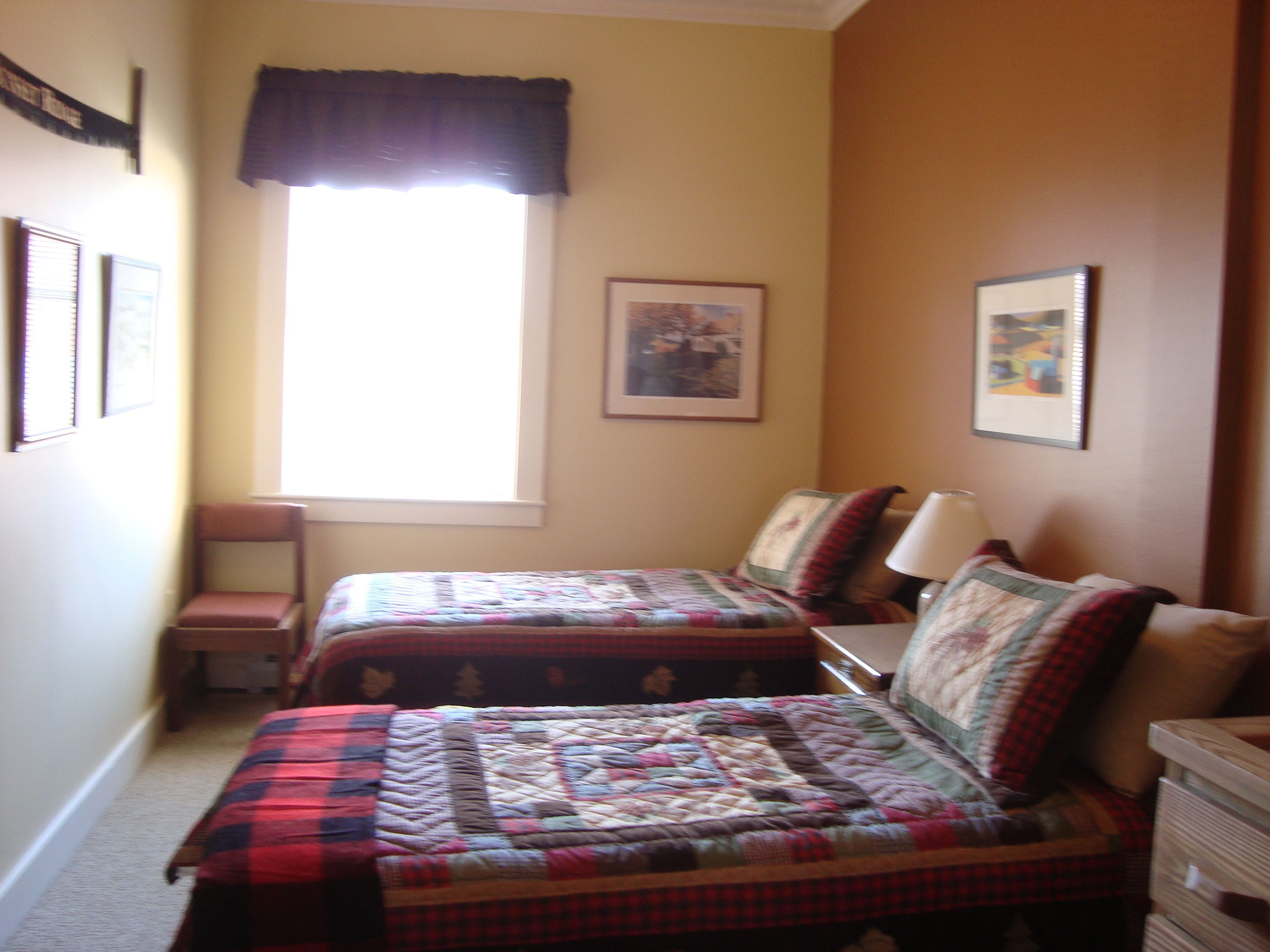 Una habitación estrecha con dos camas, una ventana con cortina oscura y cuadros en las paredes.