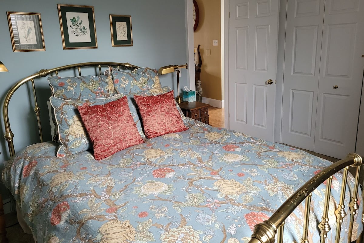 A bedroom with a bed featuring a floral patterned bedspread, a brass headboard, and two red pillows. There are framed botanical prints on the wall and a white door in the background.