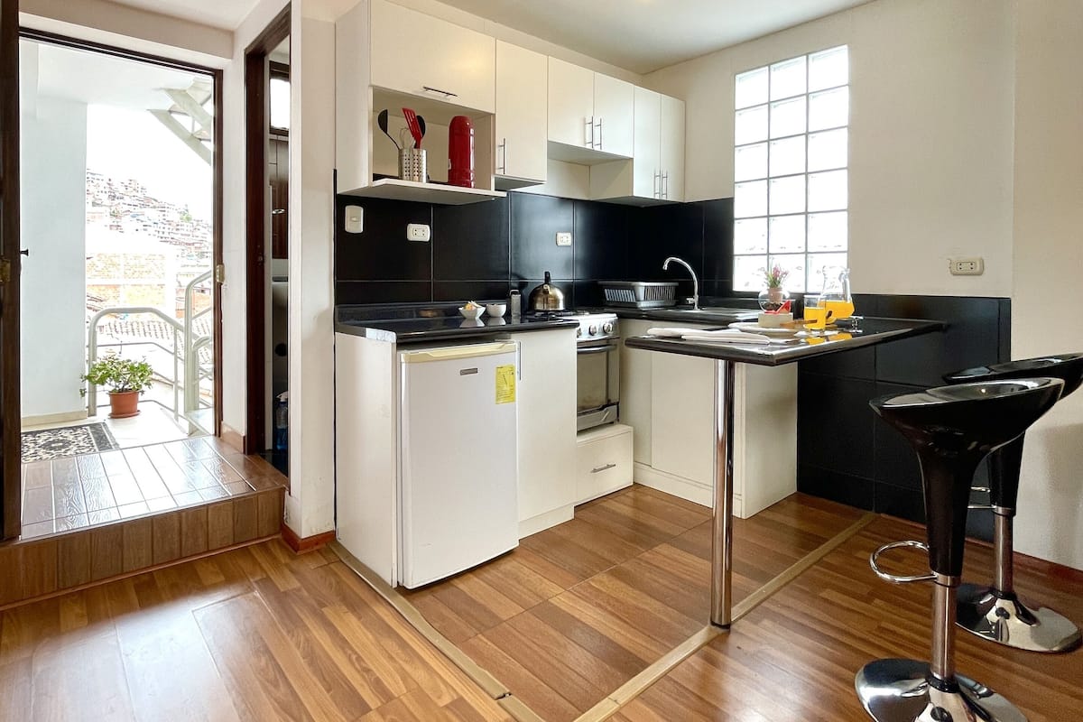 A compact kitchen with white cabinets, a black countertop, a small refrigerator, a stove, and a sink. There is a window providing natural light and a door leading outside.