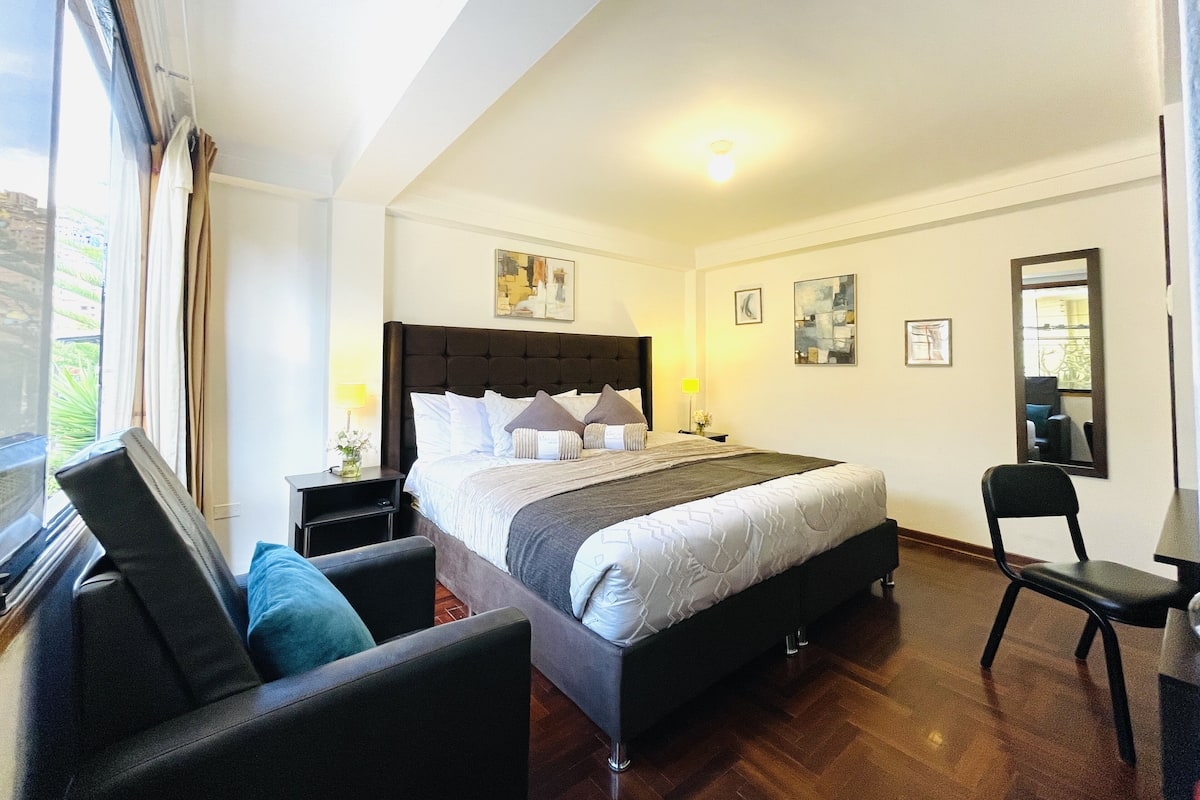 A modern bedroom with a large bed, a desk, and a chair. There is a window with curtains, a bedside table with a lamp, and artwork on the walls.