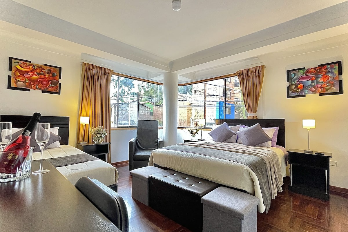 A hotel room with a bed, bedside tables, a chair, a small table with a bottle and glasses, and two windows with curtains.