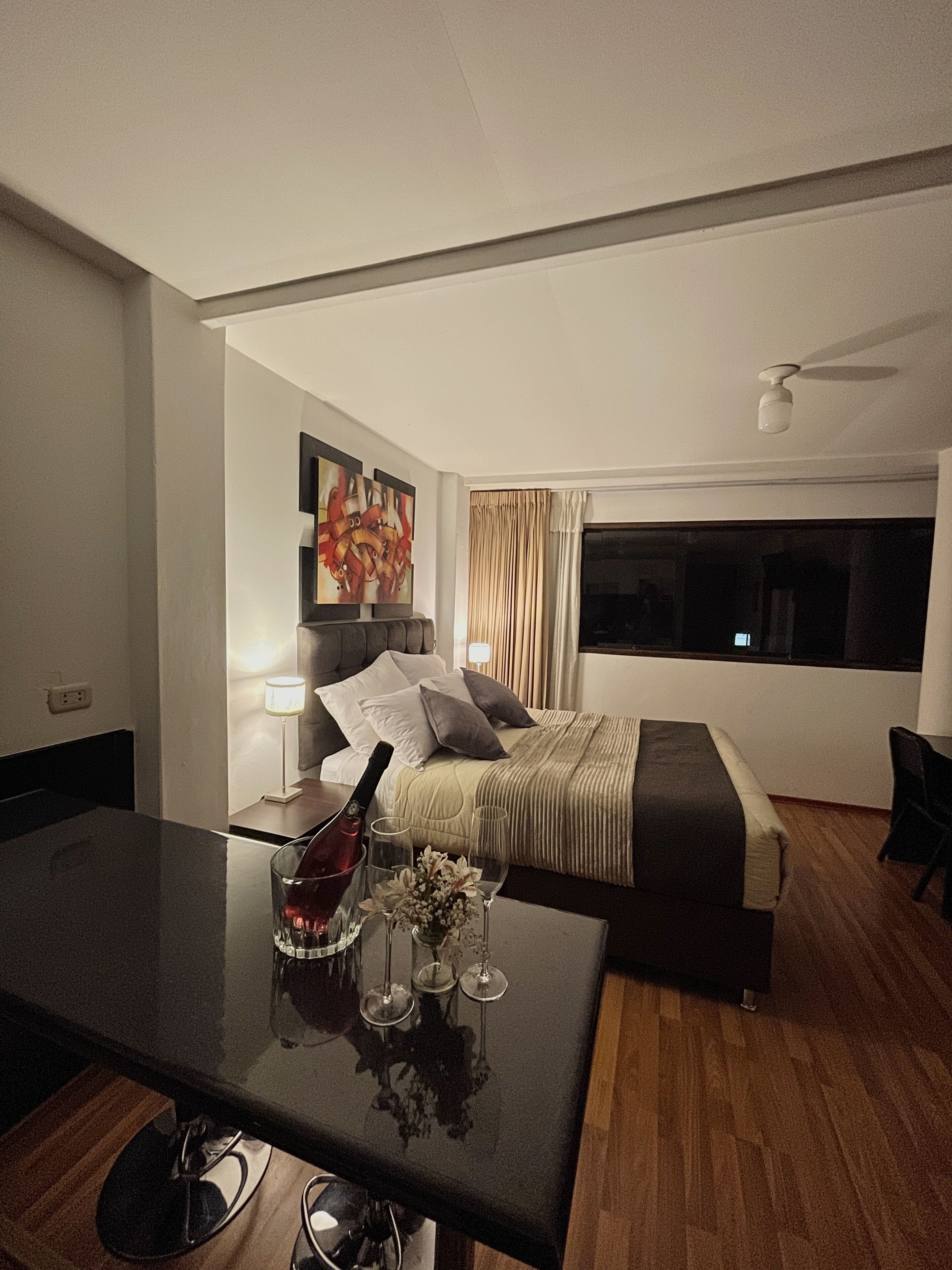 Une chambre d’hôtel avec un lit, un bureau sur lequel se trouvent une bouteille et des verres, un tableau accroché au mur et une fenêtre avec des rideaux.