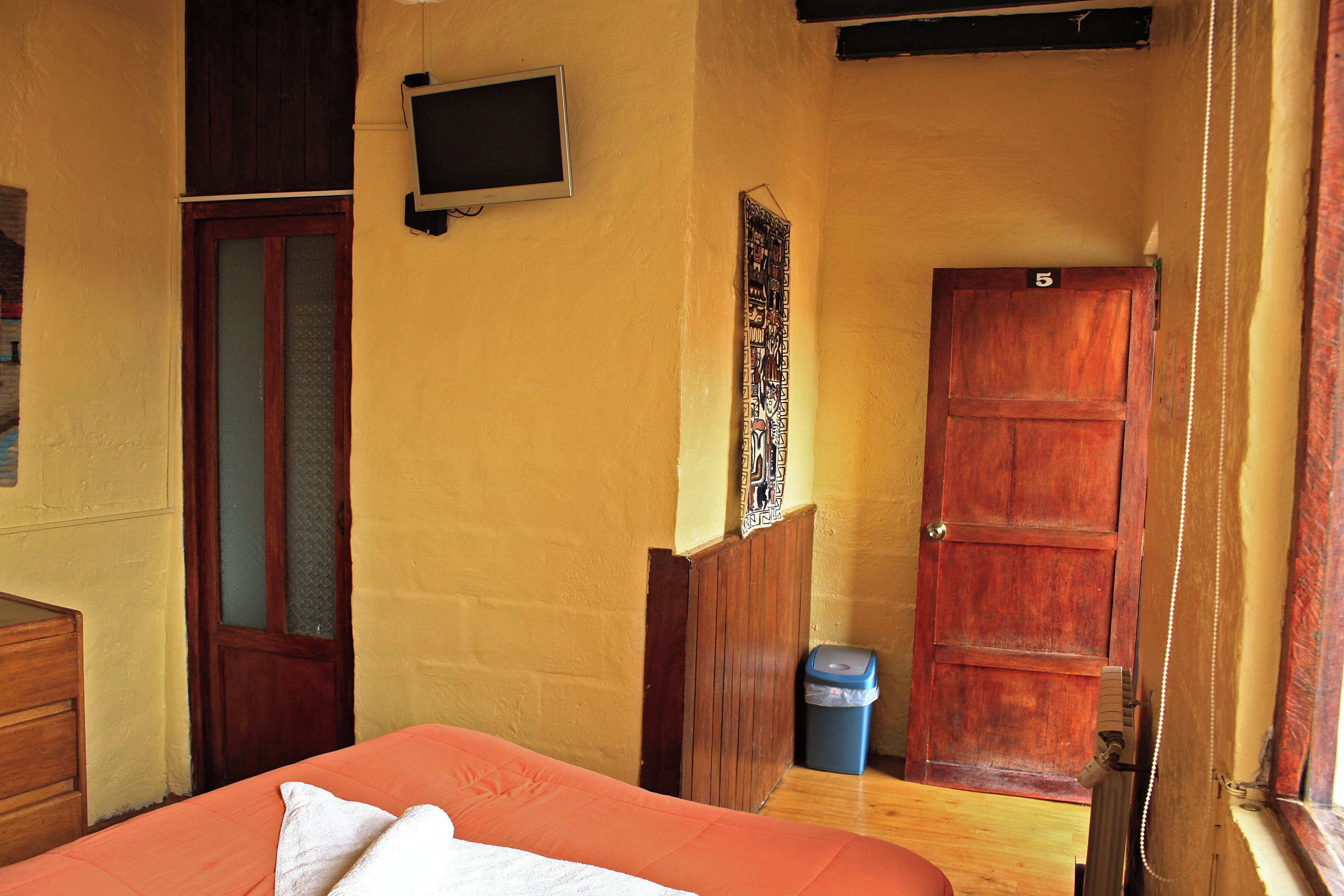 Una habitación con una cama, una puerta de madera, un cubo de basura pequeño y un televisor montado en la pared.