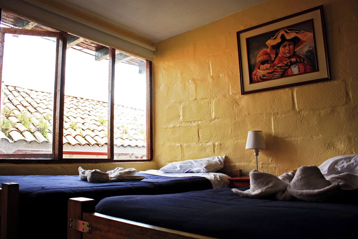 A room with two beds, a window with a view of a tiled roof, a framed painting on the wall, and a bedside lamp.