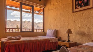Una habitación con dos camas, techo de madera, ventana con vista a la montaña, lámpara de pared e imagen en un marco.