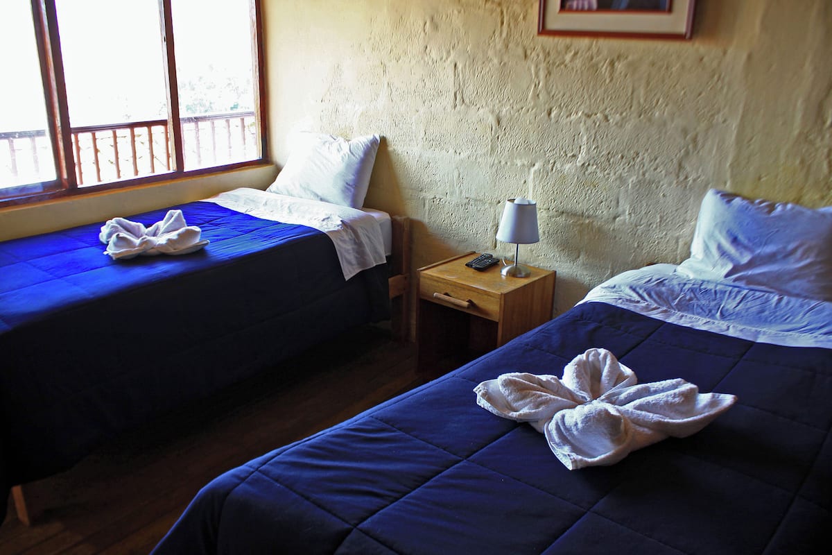 A room with two beds, each with a blue bedspread and white pillows. There is a wooden nightstand with a lamp and a remote control.