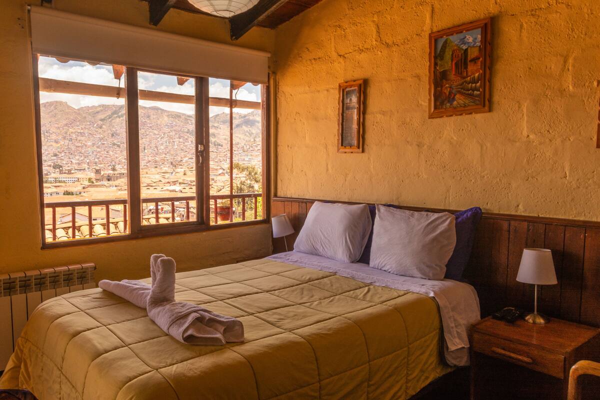 A hotel room with a bed, a nightstand, a window with a view of a cityscape, and a painting on the wall.