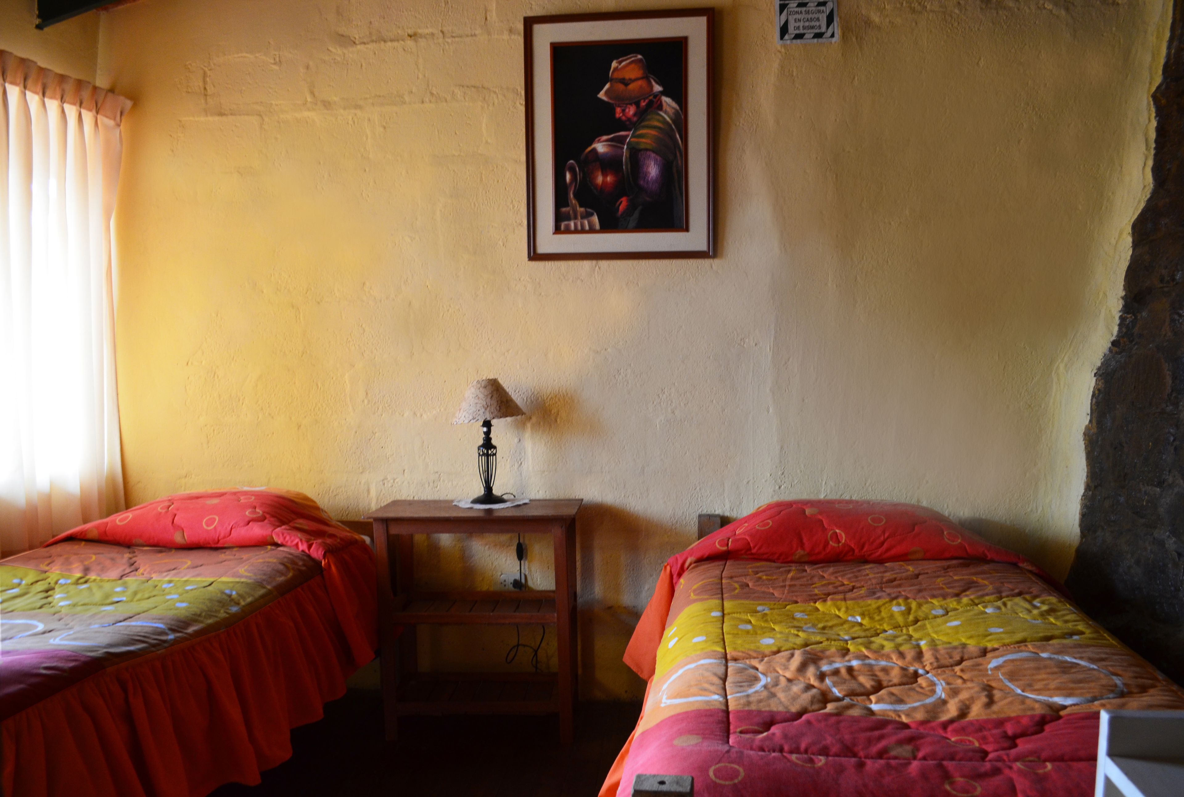 A room with two single beds, a small table with a lamp, a framed picture on the wall, and a window with curtains.