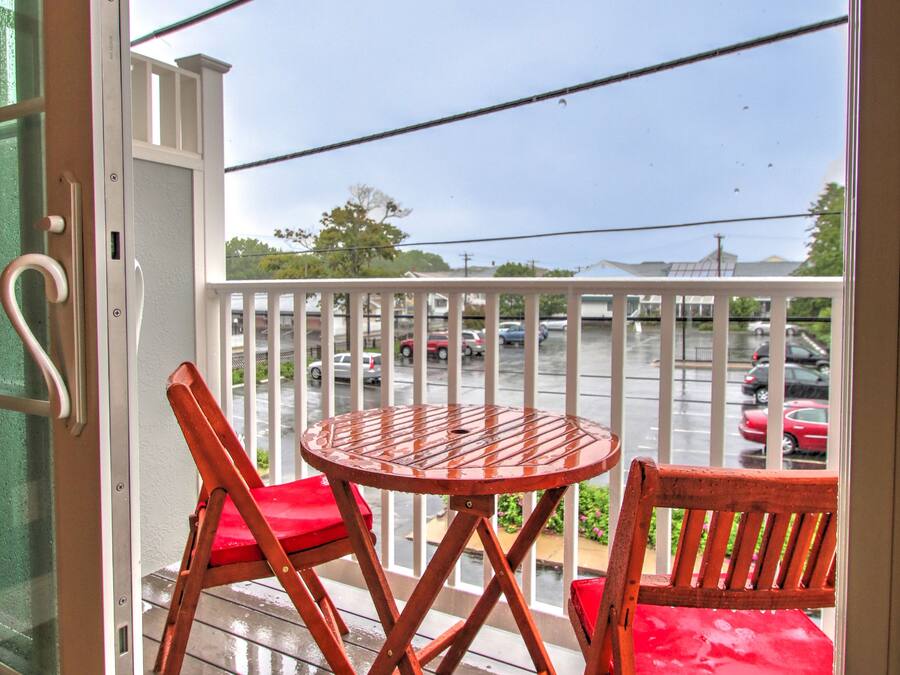 Una terraza de madera con una mesa y sillas, barandilla blanca, y vista a un estacionamiento.